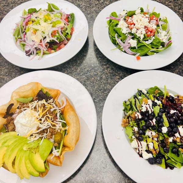 Four diverse salads, including a taco salad and a blueberry and goat cheese salad, on a dark surface.